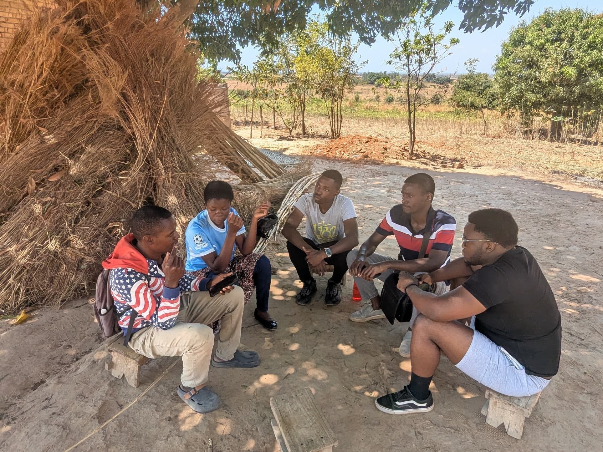 Five people sit on low wooden stools in the shade of a tree. One holds a phone and explains while the others listen.