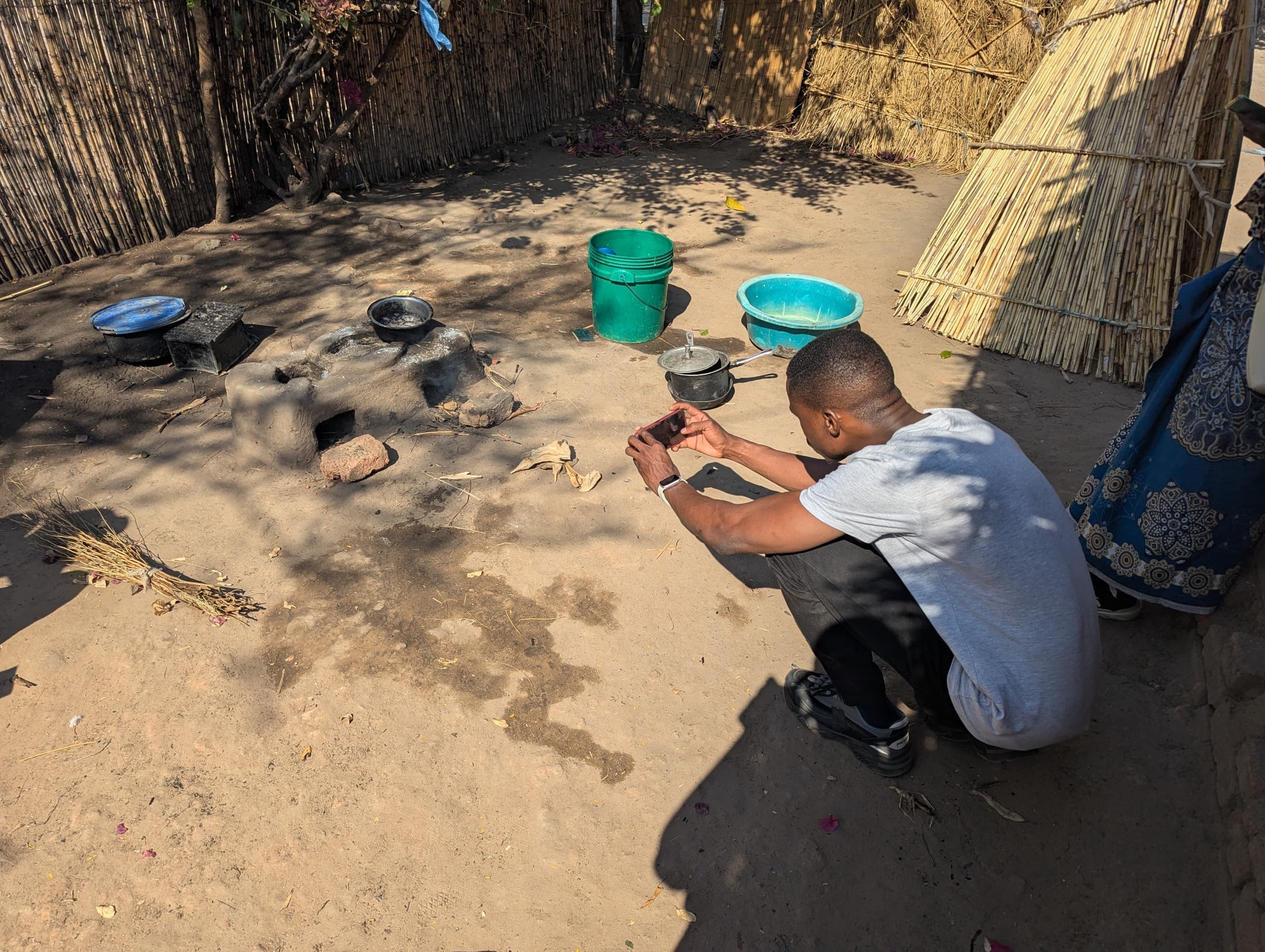 Crouched in a reed-fenced cooking area, photographing a clay Changu Changu Moto stove with a phone during an inspection.