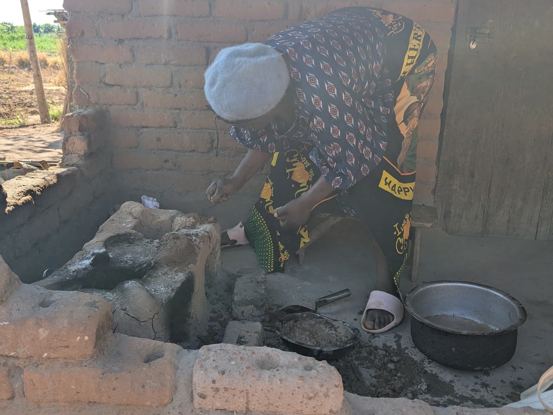 A woman in a grey knitted hat bends over a clay Changu Changu Moto, working fresh mud into it by hand, with a pan of mud and a cooking pot beside her.
